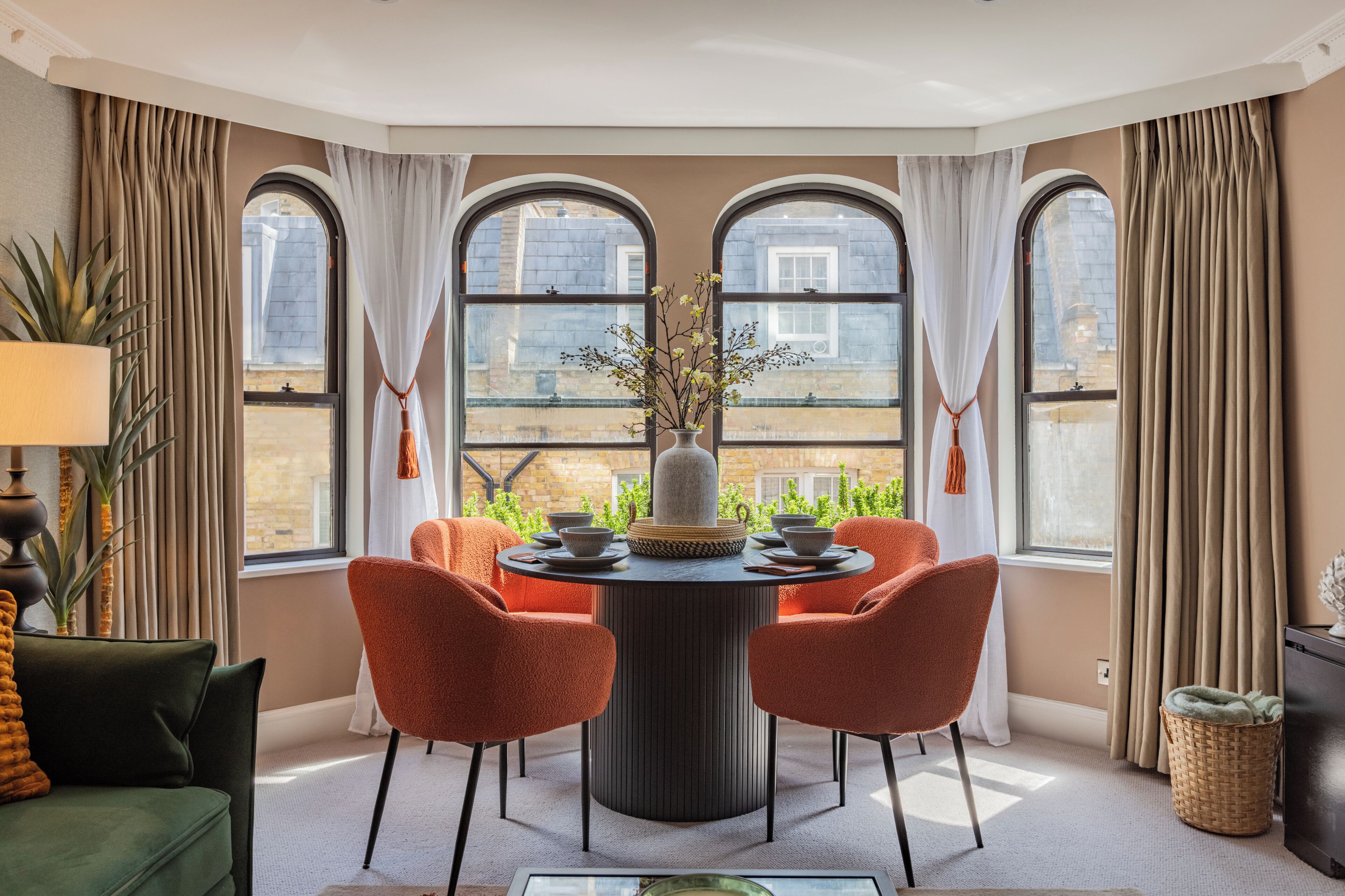 A room with a round table, four chairs, a vase with flowers, and large windows with curtains.