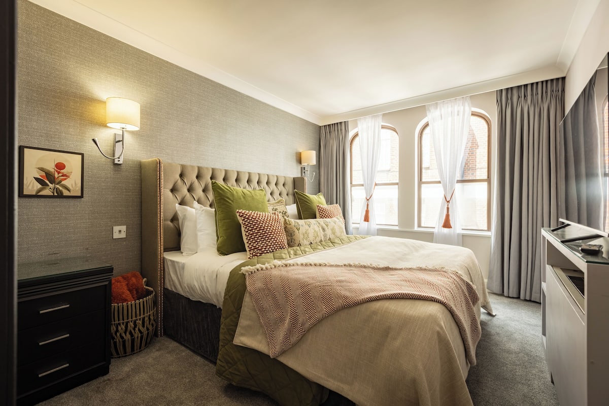 A bedroom with a tufted headboard, a bed with multiple pillows, a bedside table, a lamp, and a window with curtains.