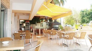An outdoor cafe with wicker tables and chairs, a bar area, and a yellow striped awning. - Nasa Flora Hotel (Antalya)