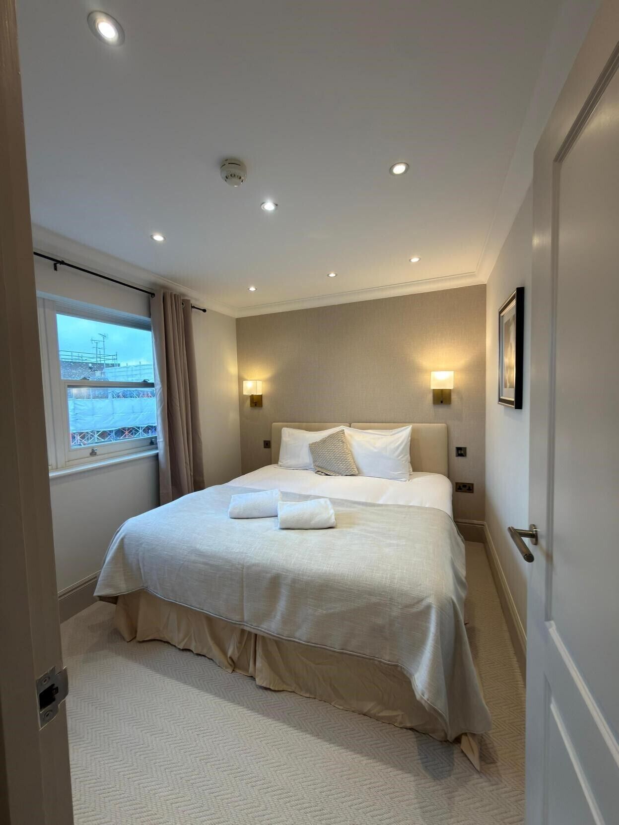 A hotel room with a bed, two bedside lamps, a window with curtains, and a view of a building outside.