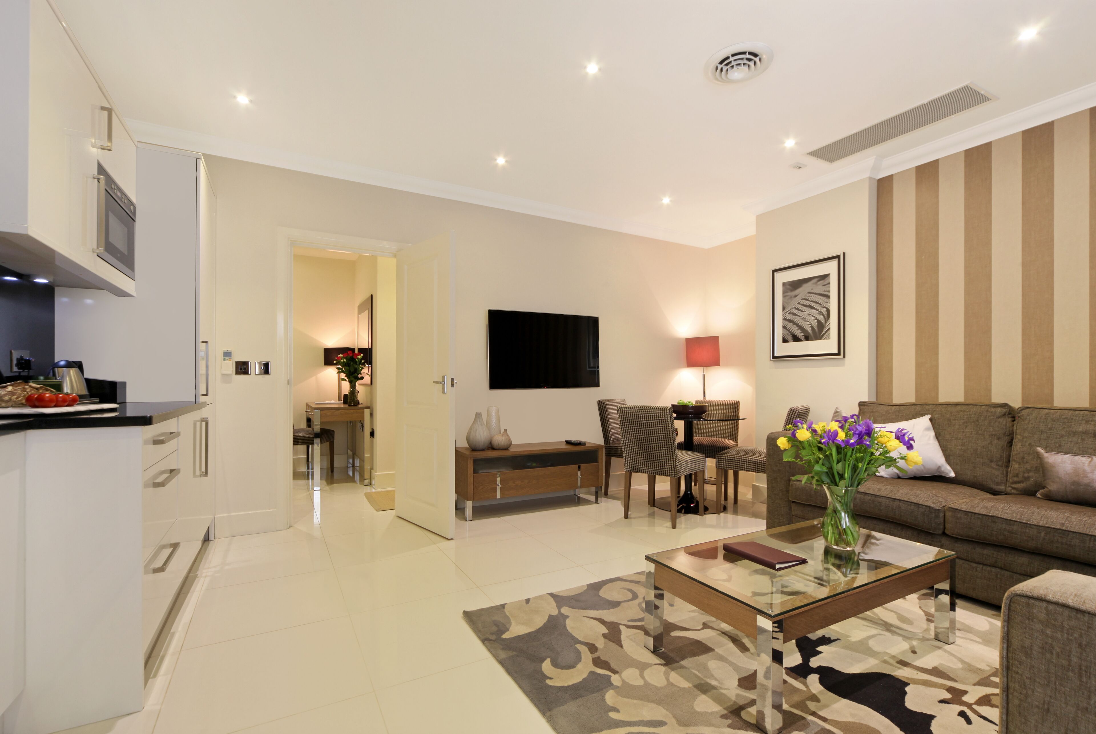 A modern living room with a sofa, coffee table, and TV. There is a kitchen area with cabinets and a dining table.