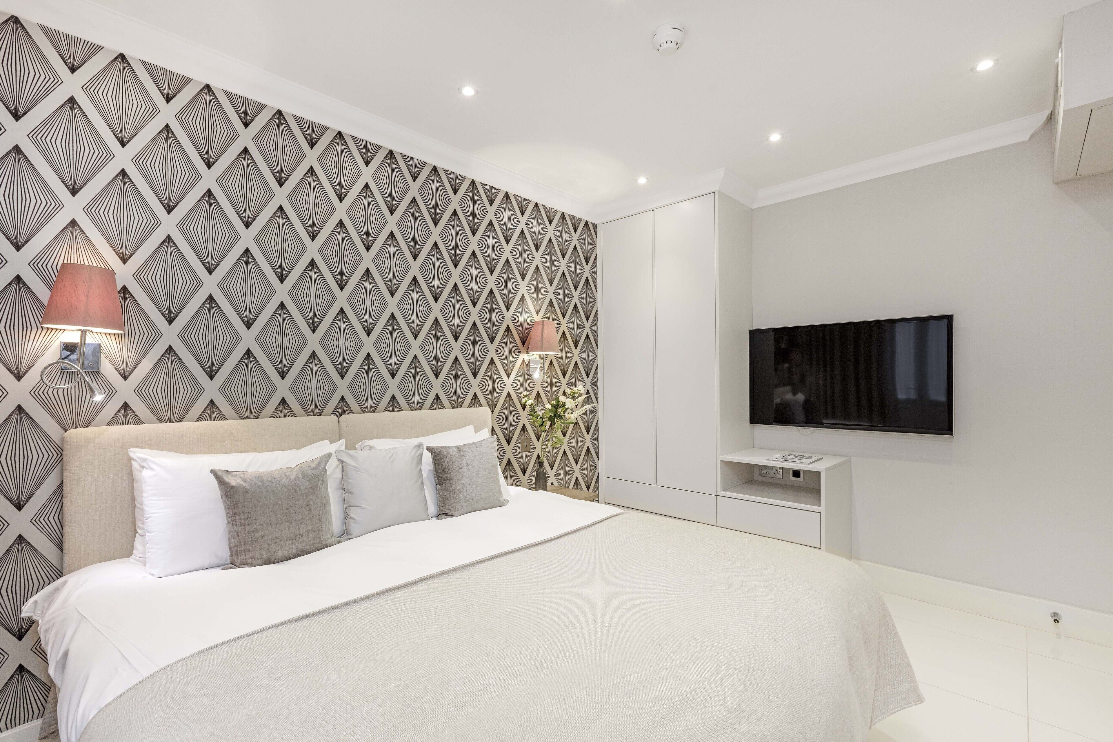 Une chambre à coucher moderne avec un papier peint à motifs, un grand lit avec une literie blanche, deux lampes de chevet, un téléviseur à écran plat et une petite table de chevet.