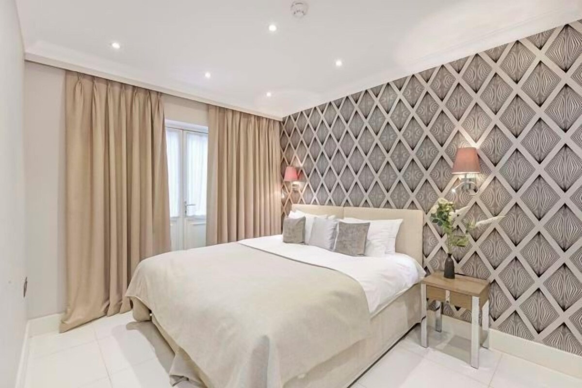 A neatly arranged bedroom with a bed, curtains, a nightstand, and a wall with a geometric pattern.