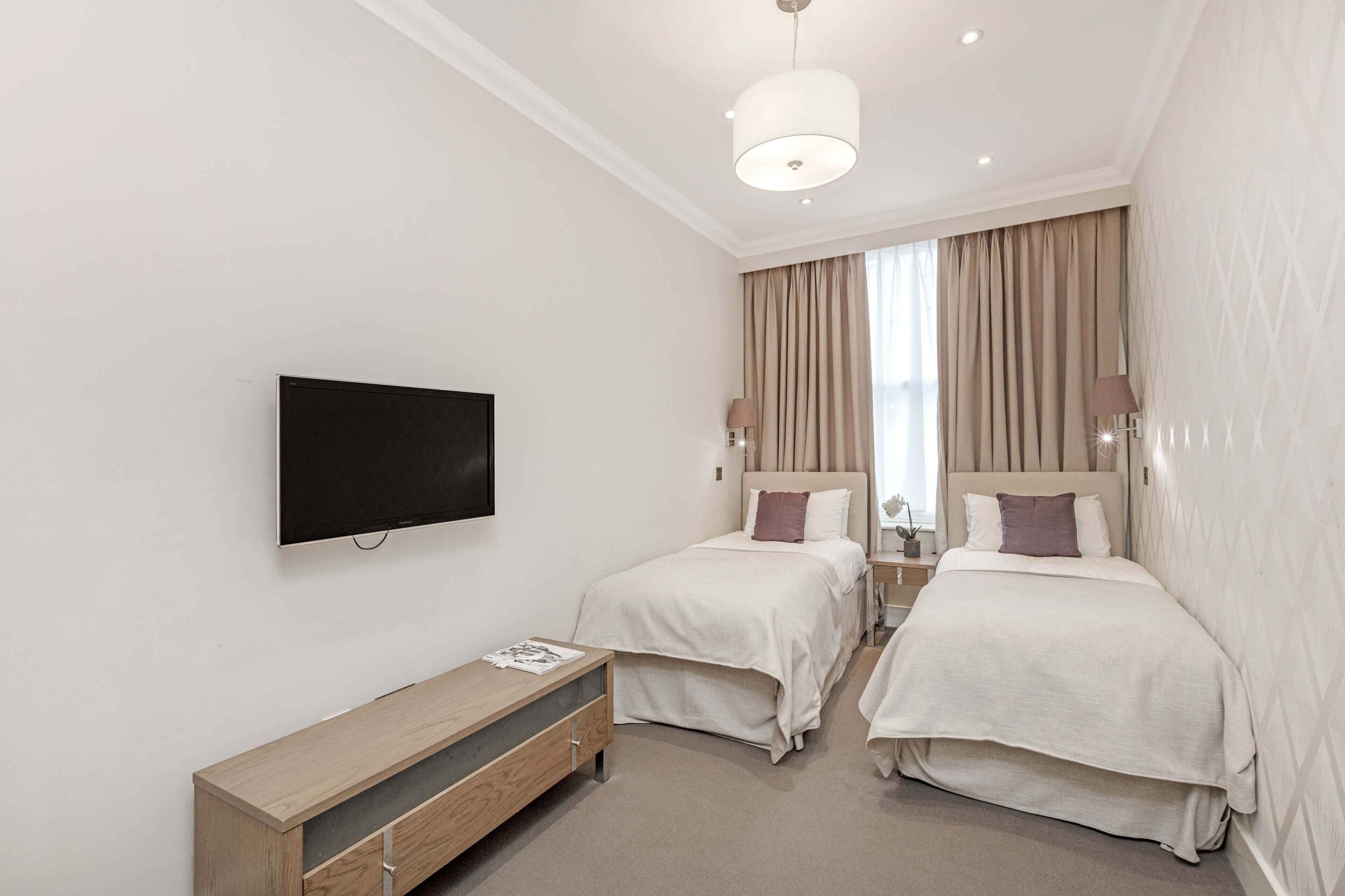 A hotel room with two beds, a TV, a wooden nightstand, and a large window with curtains.