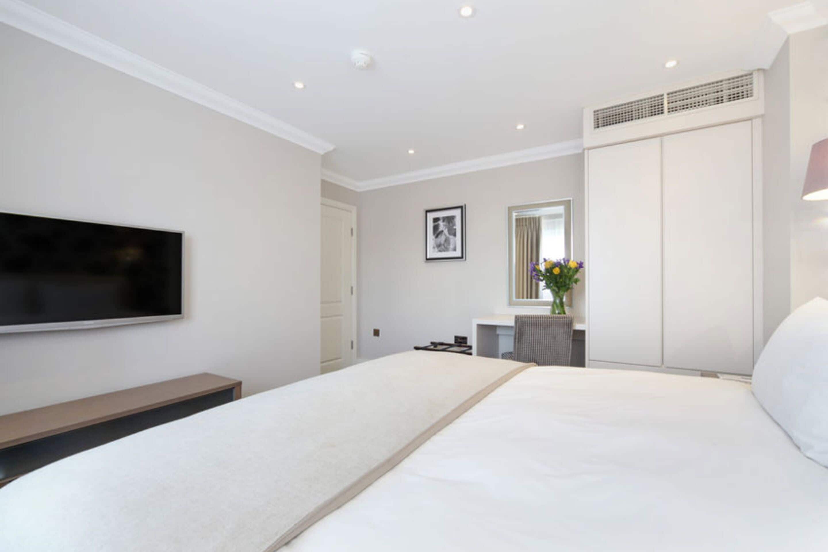 A modern hotel room with a large bed, a flat-screen TV mounted on the wall, a bedside table, a mirror, and a wardrobe.