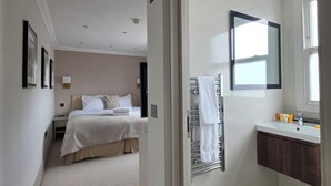 Una camera d'albergo con un letto, un bagno con lavandino e specchio, e un corridoio che porta a un'altra stanza.