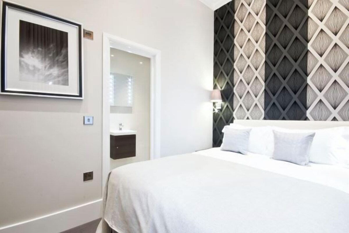 A hotel room with a bed, a framed picture on the wall, a bathroom with a sink, and a patterned wall.