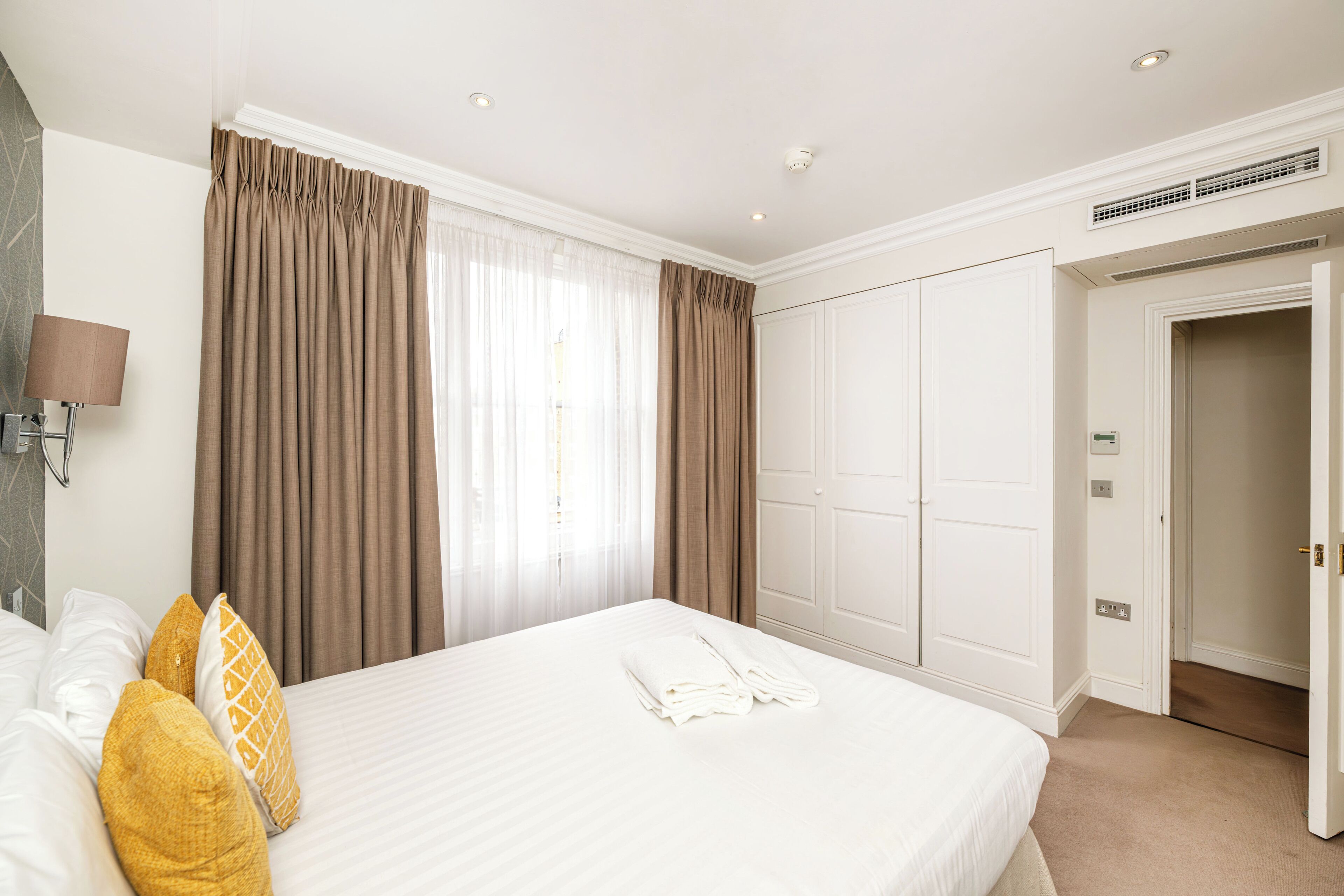 A hotel room with a bed, curtains, a wardrobe, and a door leading to another room.