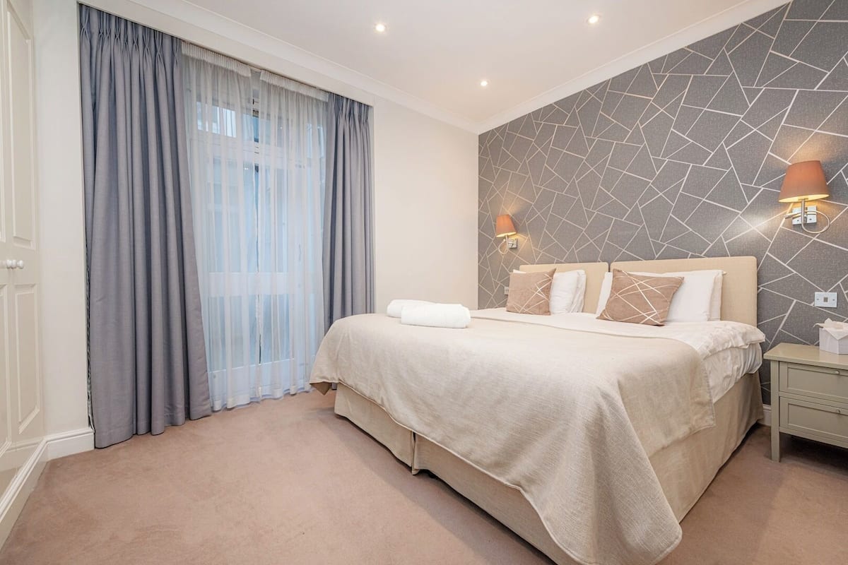 Quarto de hotel com cama, cortinas, uma parede com padrão geométrico e mesa de cabeceira com abajur.