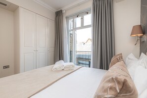 Un lit bien fait, avec une tête de lit en bois, une pile de serviettes pliées, et une vue sur un balcon à travers une fenêtre ornée de rideaux gris.