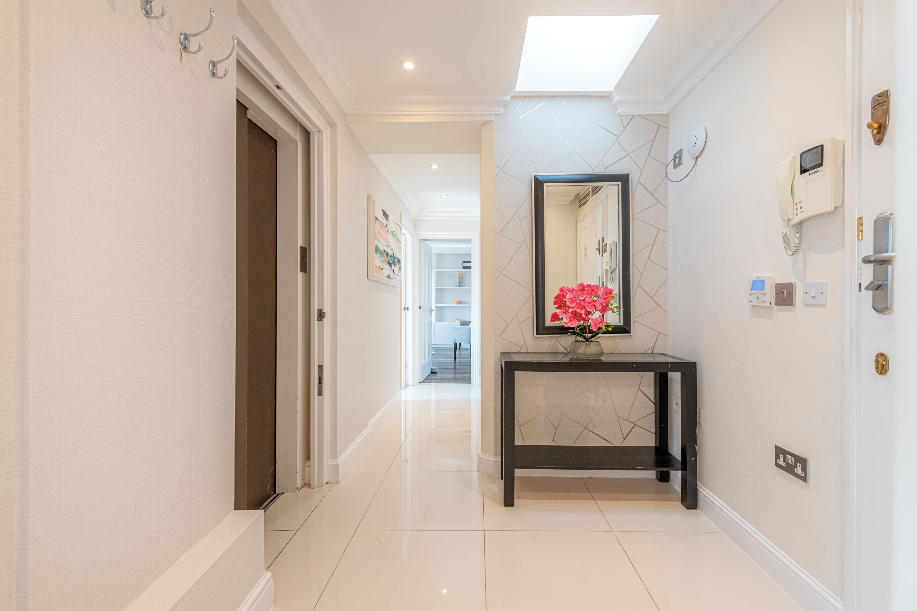 A modern hallway with a glass door, a console table with a vase of flowers, a wall-mounted phone, and a light fixture.