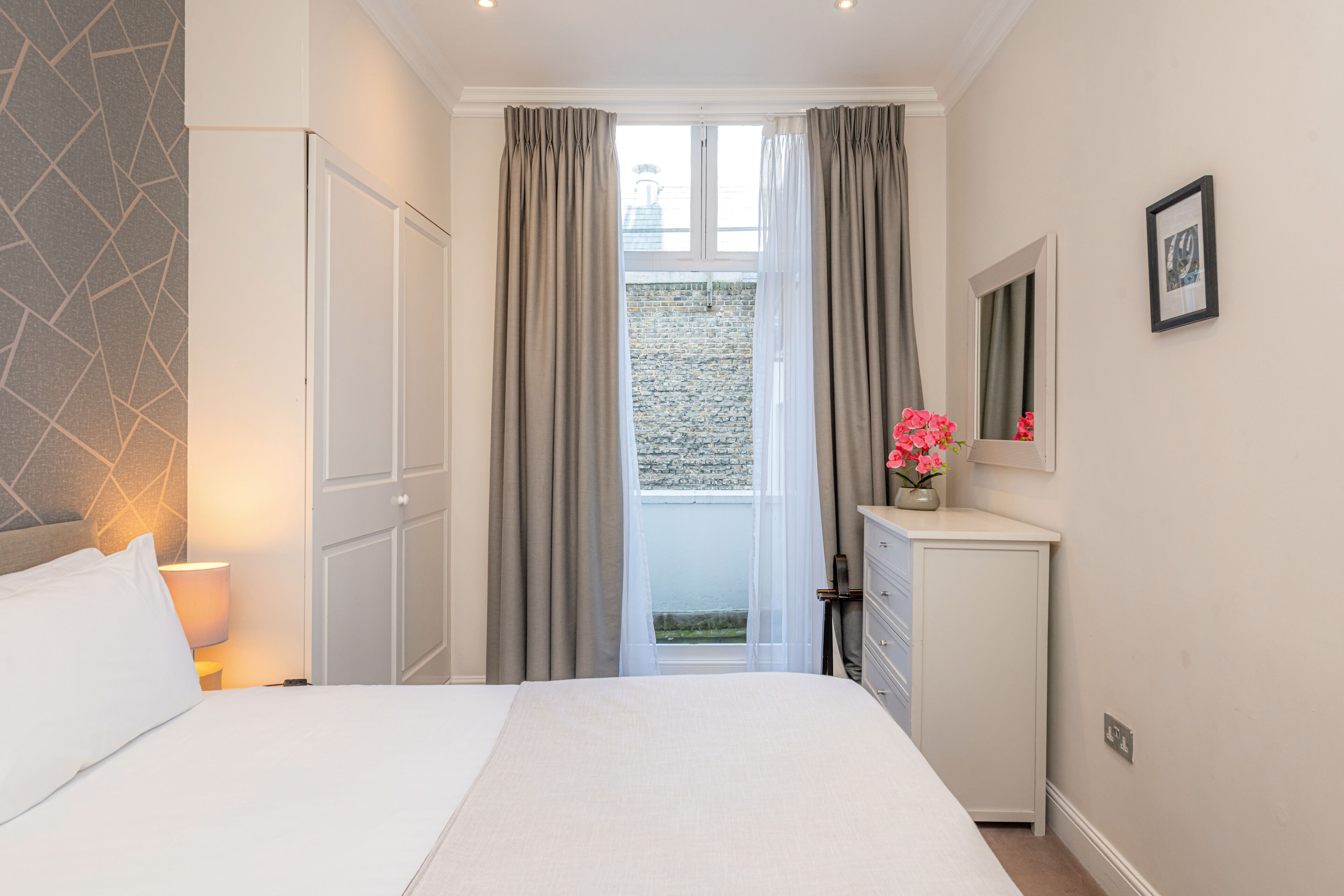 Ein Schlafzimmer mit Bett, Schminktisch, Spiegel, Fenster mit Vorhängen und Blick nach draußen.