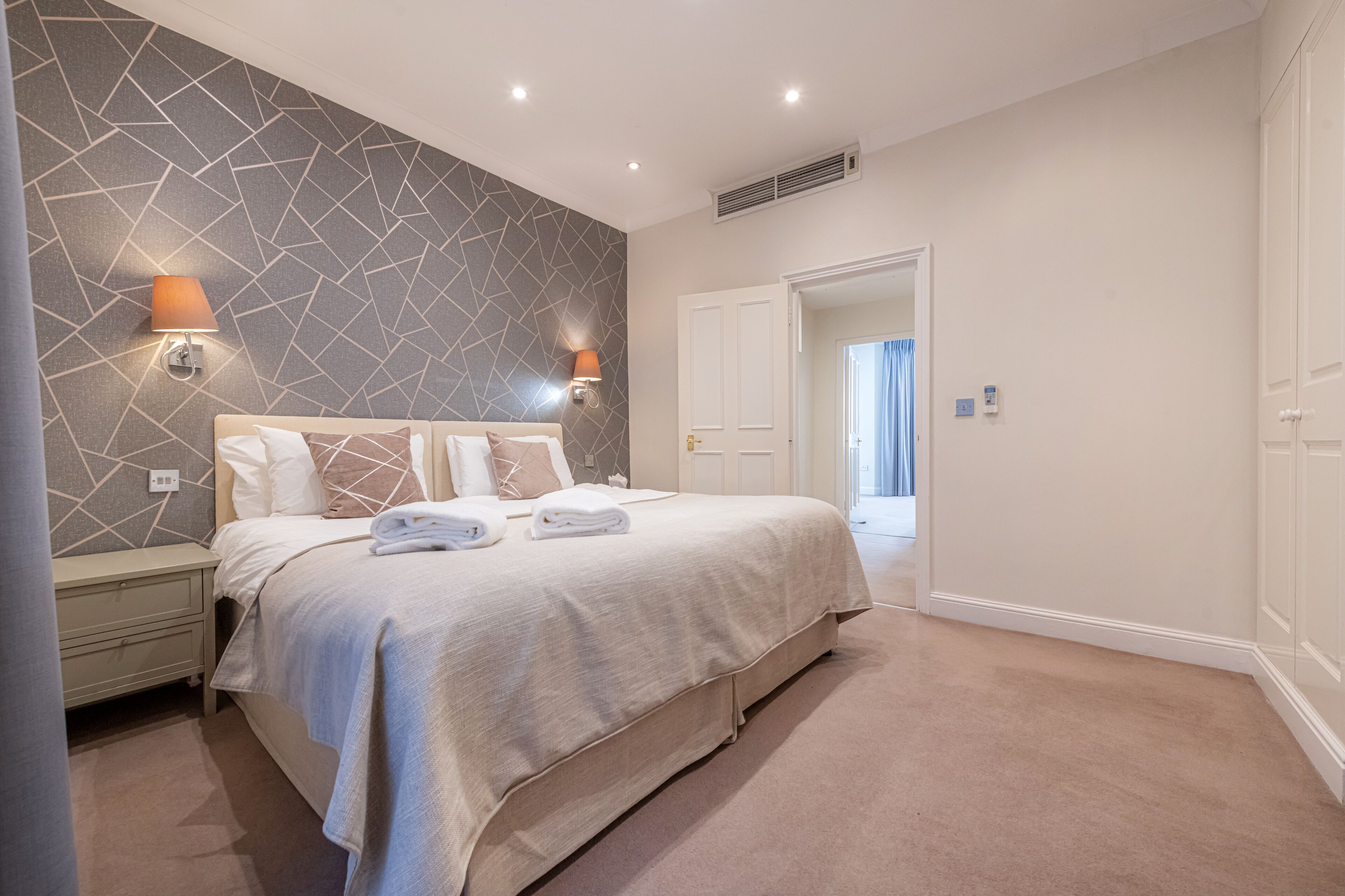 A modern bedroom with a geometric-patterned wall, a large bed with a beige bedspread, two wall-mounted lamps, and a door leading to another room.