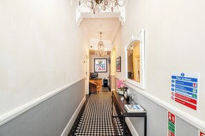 Un couloir d’hôtel avec un sol à carreaux, un lustre, un miroir et un panneau indiquant les consignes de sécurité incendie.