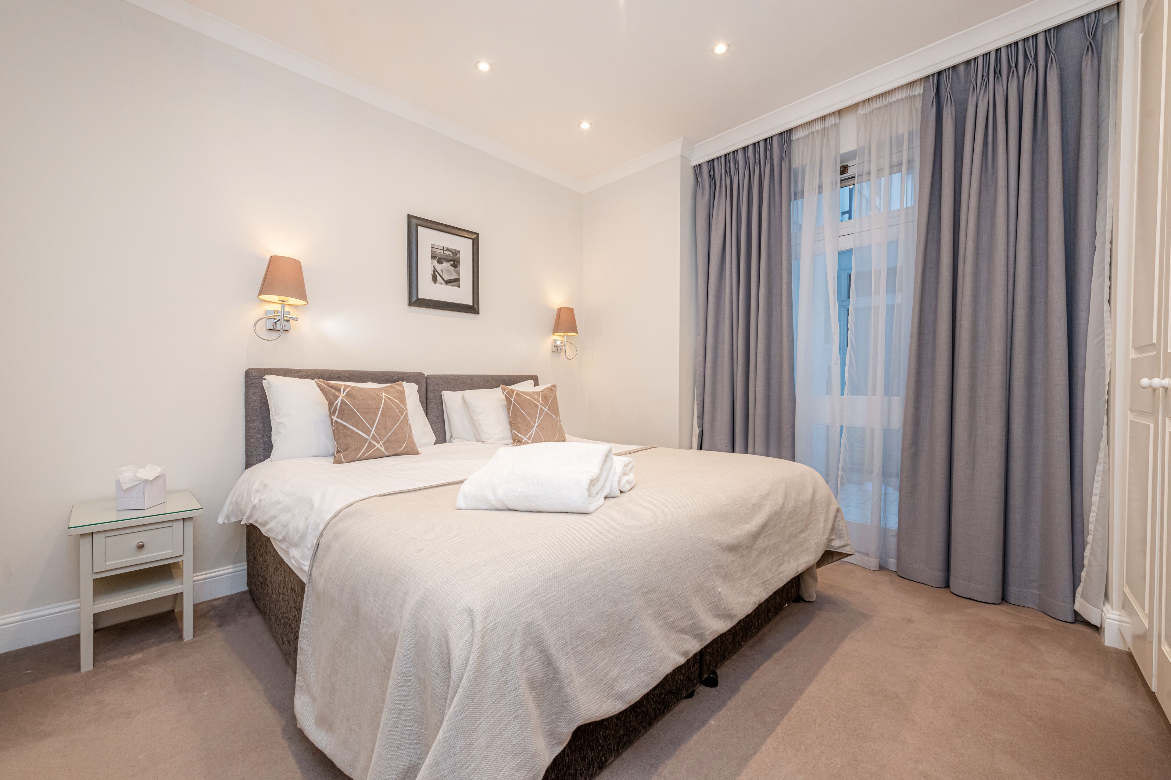 A neatly made bed with a white comforter and pillows, a small bedside table with a tissue box, and a wall-mounted picture.