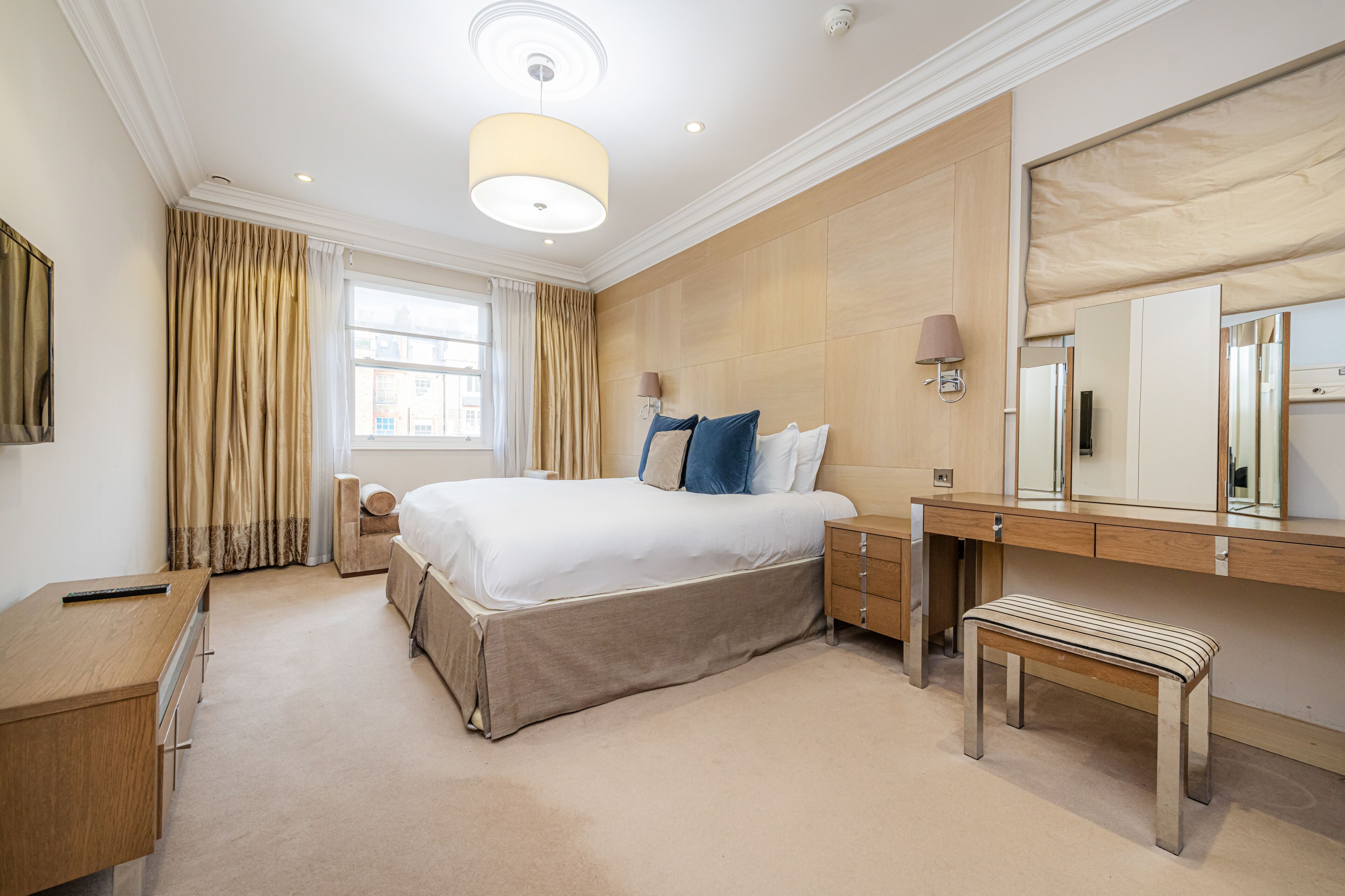 Une chambre d’hôtel avec un grand lit, un bureau en bois, un banc, une commode et un grand miroir.