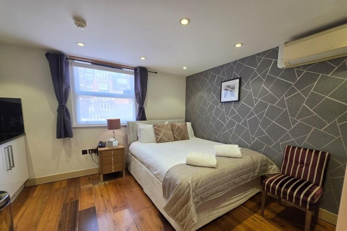 Ein Hotelzimmer mit einem Bett, einem Sessel, einem kleinen Tisch mit Lampe, einem Fenster mit Vorhängen und einem Wandbild.