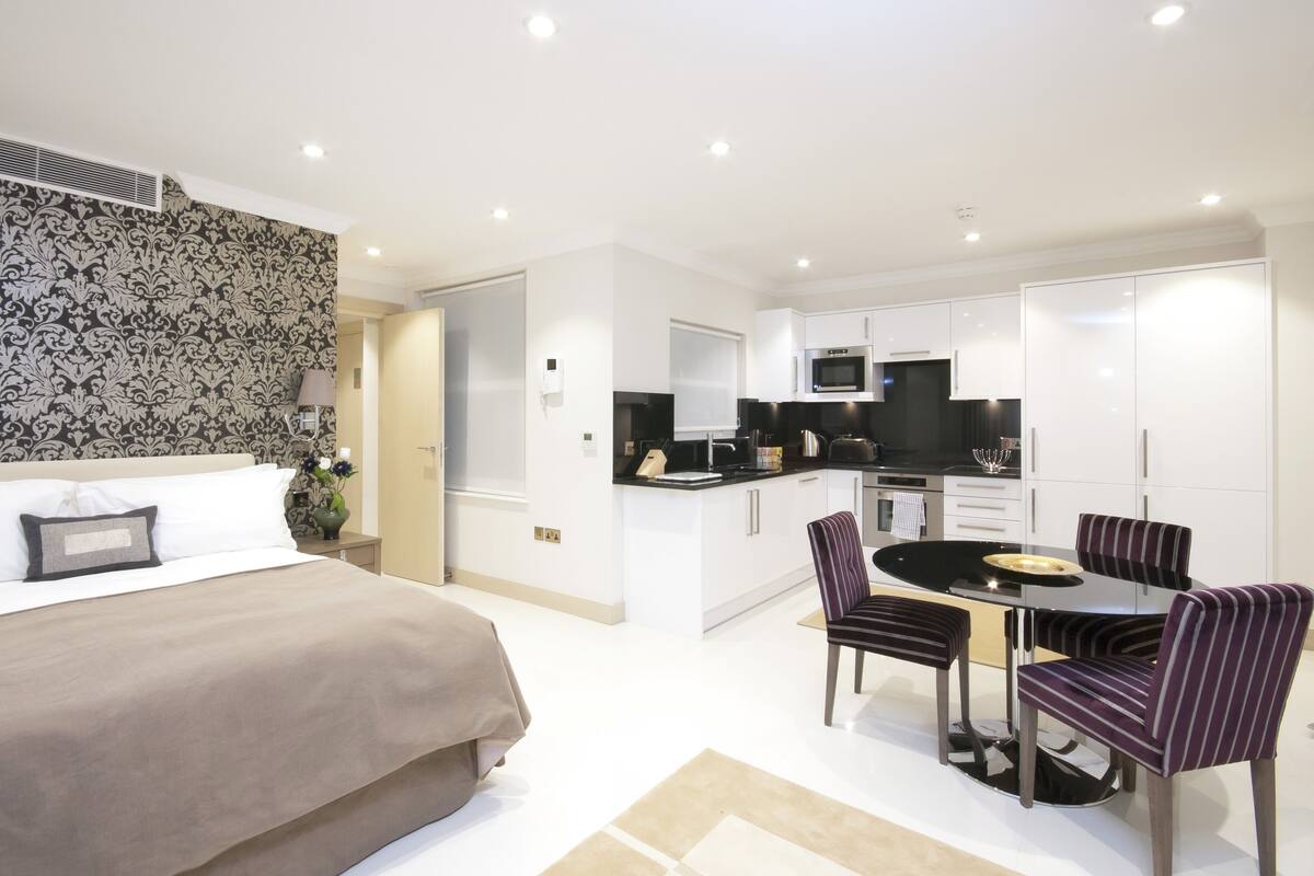 A modern hotel room with a bed, a dining area with a table and chairs, a kitchen area with a microwave and cabinets, and a patterned wallpaper on the wall.