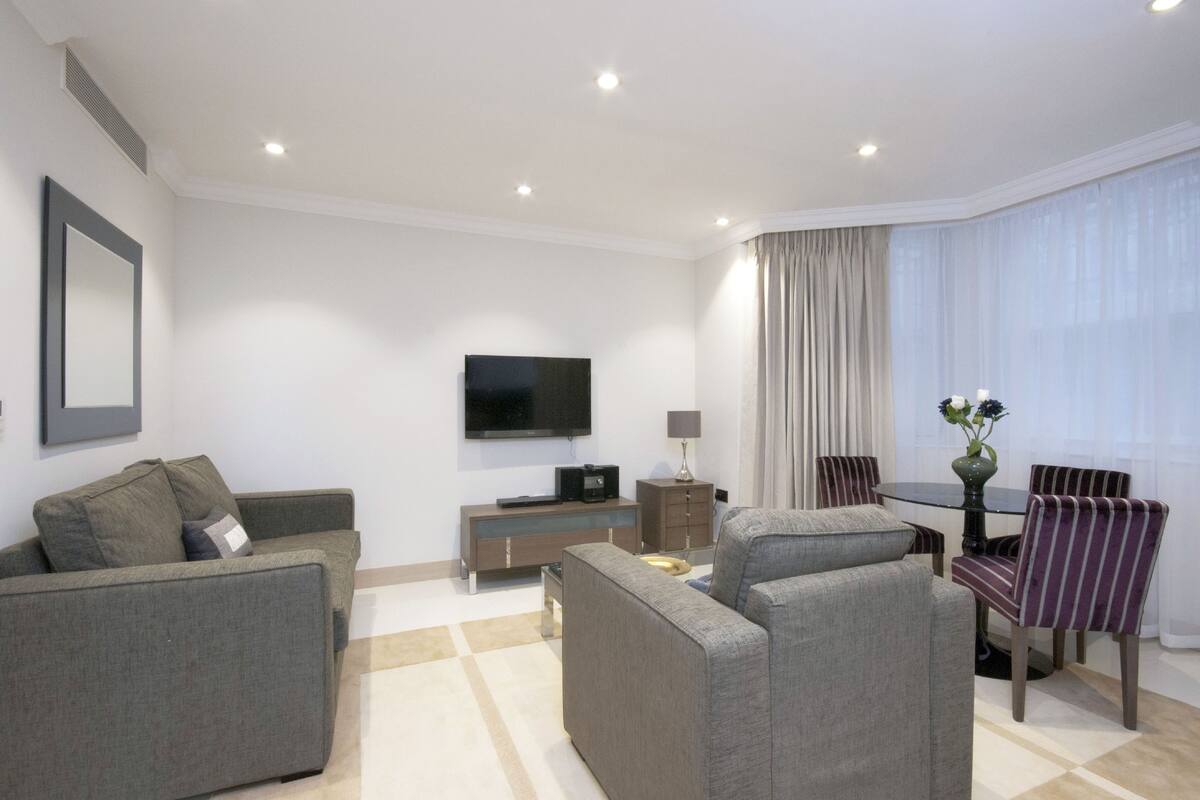 A modern living room with a flat-screen TV, grey sofas, a dining table with chairs, and a large window with curtains.