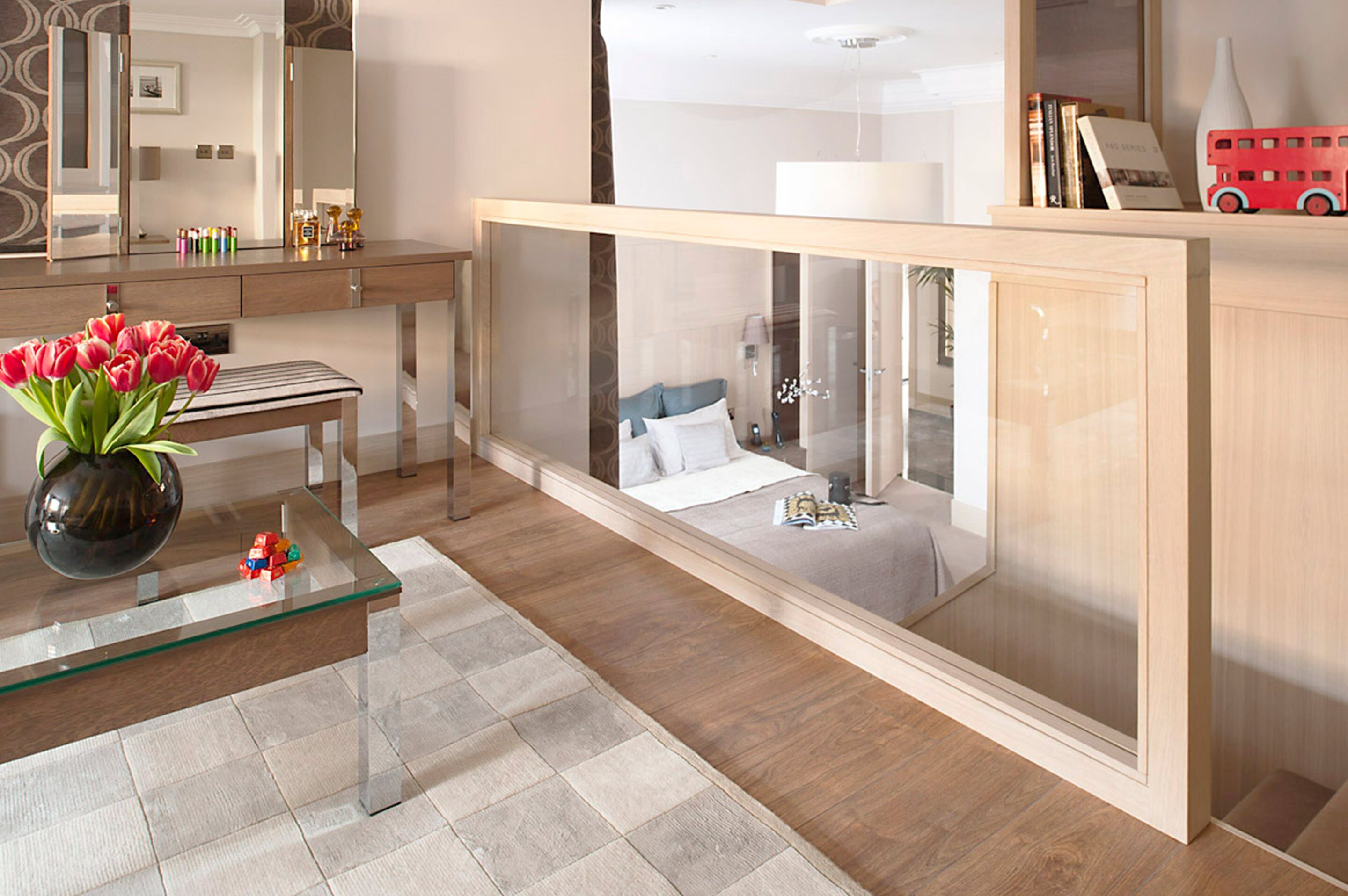 A modern hotel room with a glass partition, a wooden vanity, a bed with a blue headboard, and a red toy bus on the shelf.