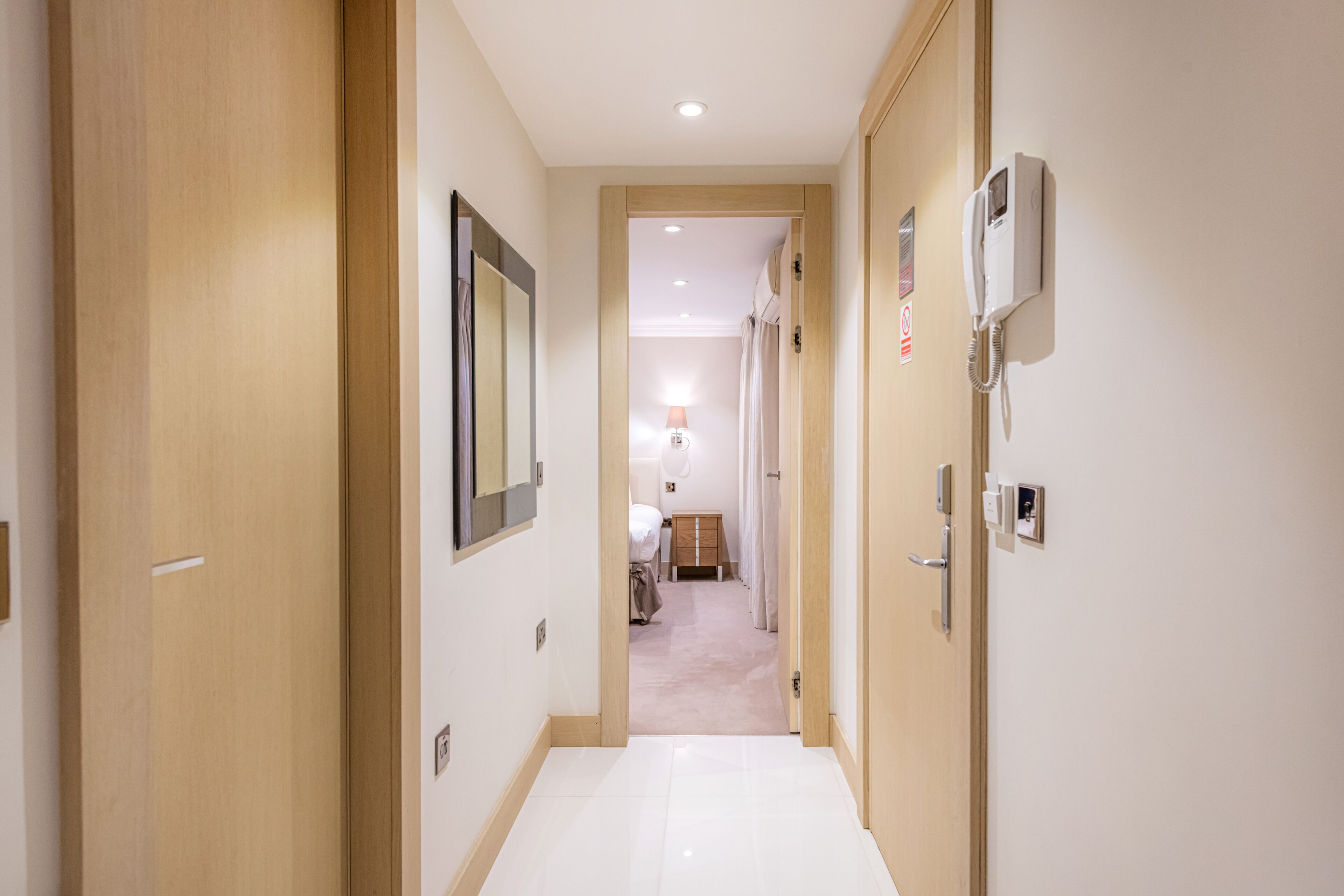 Een hotelgang met houten deuren, een vloer van witte tegels en een bed in de verte.