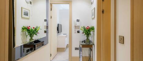 A hallway with a mirror, a small table with a vase of flowers, and a door leading to a room.