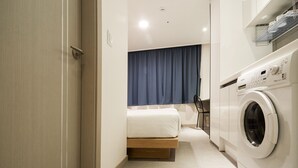 Habitación de hotel compacta con cama, lavadora y un pequeño rincón de descanso.