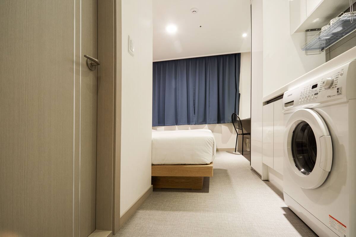 Habitación de hotel compacta con cama, lavadora y un pequeño rincón de descanso.