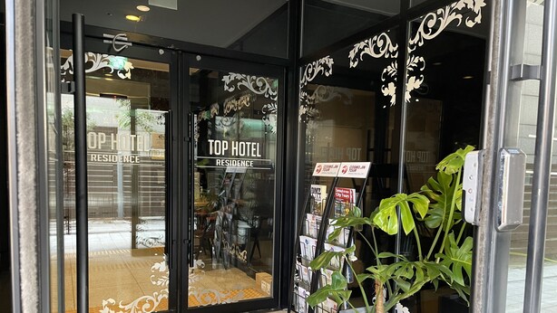 La entrada de un hotel con puertas de cristal, un letrero que indica 'Top Hotel Residence', y una planta afuera.