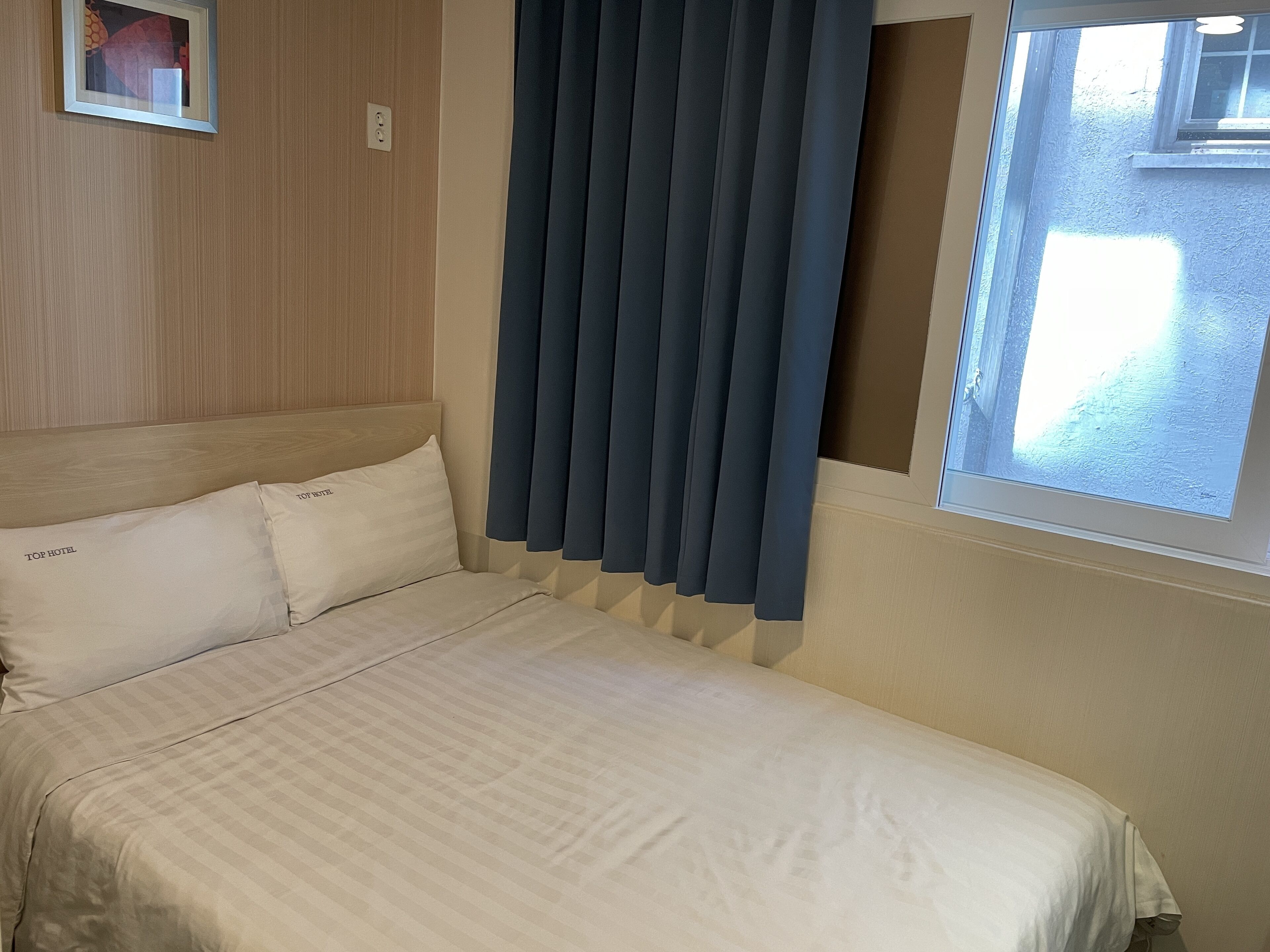 A hotel room with a bed, two pillows, a window with curtains, and a wall-mounted light switch.