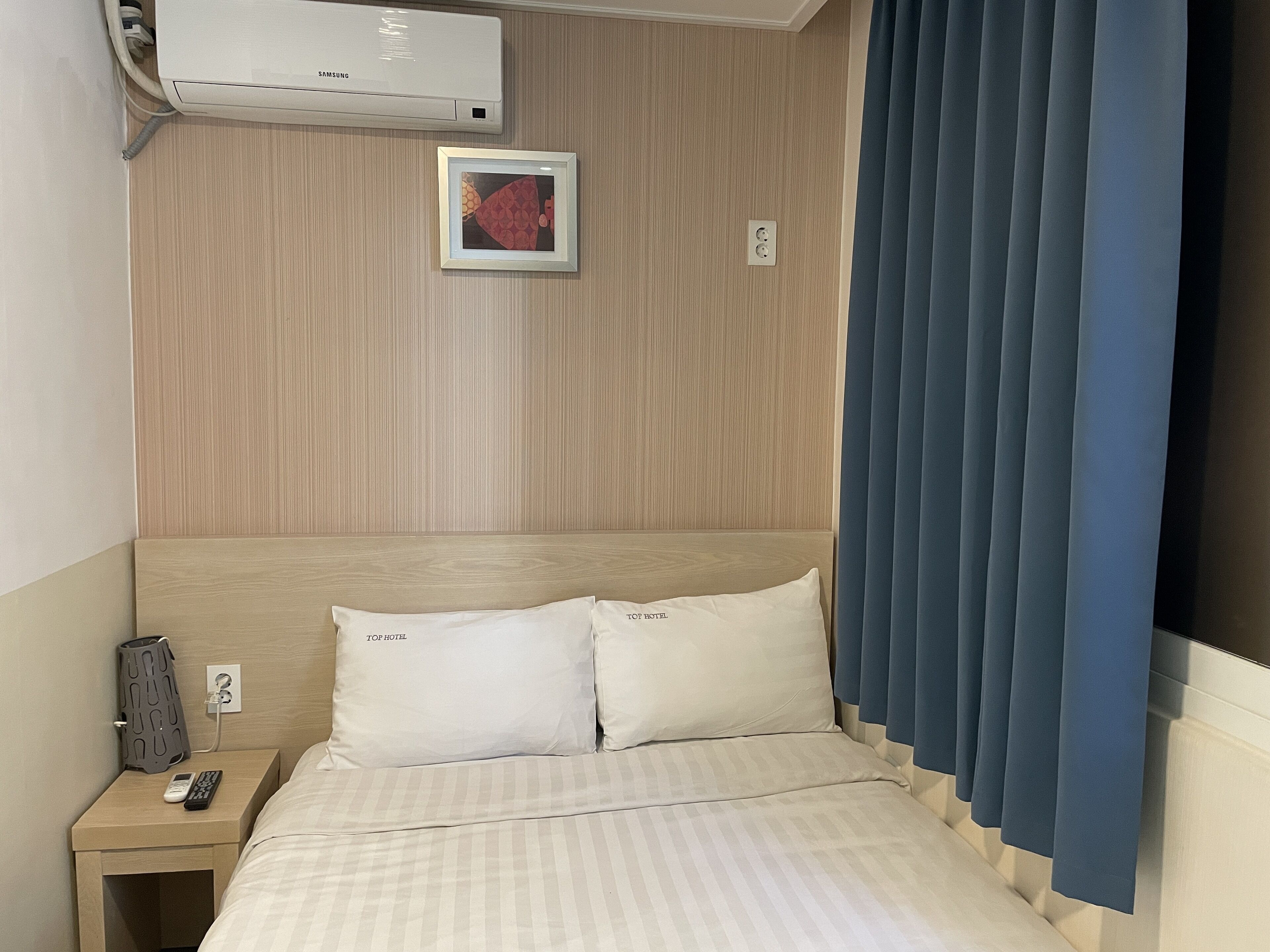 Ein Hotelzimmer mit einem Bett, einem Nachttisch, einer Fernbedienung, einer Wand-Klimaanlage und einem gerahmten Bild.