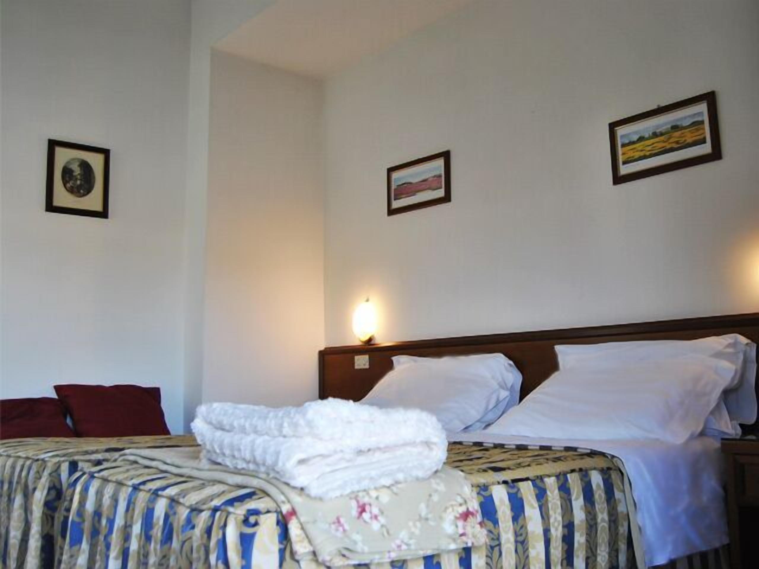 Ein Hotelzimmer mit zwei Betten, jedes mit einer anderen Musterdecke. An den Wänden hängen zwei gerahmte Bilder, eines zeigt eine Landschaft, das andere einen Kreis.