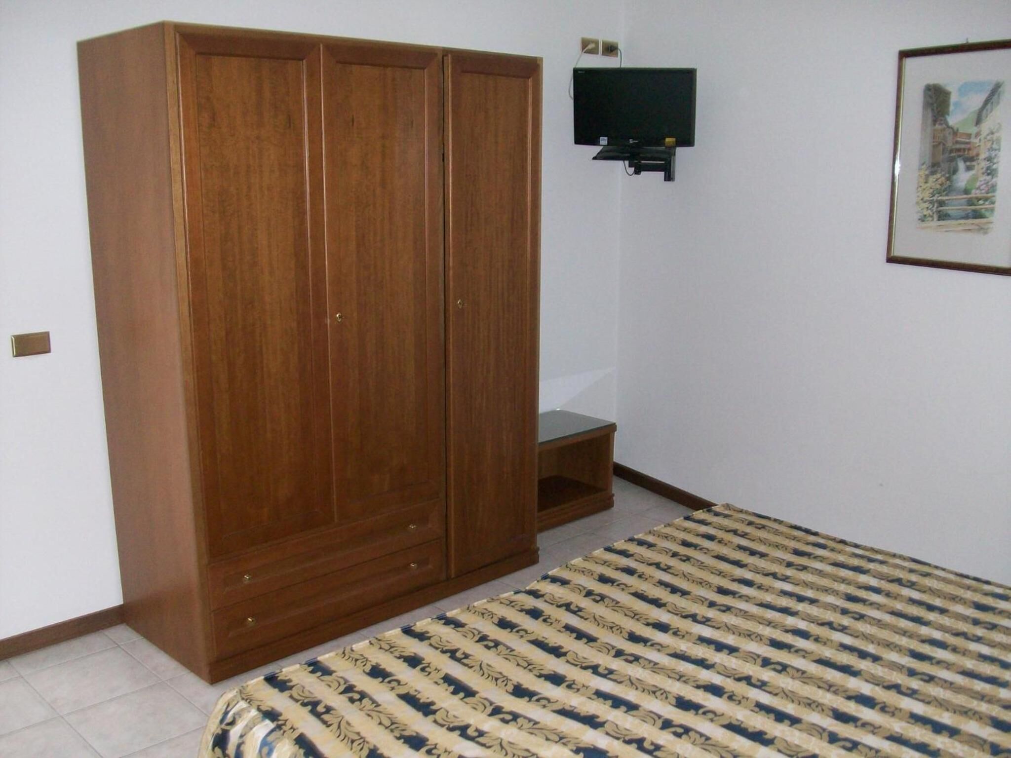A room with a wooden wardrobe, a small TV mounted on the wall, and a patterned bedspread.