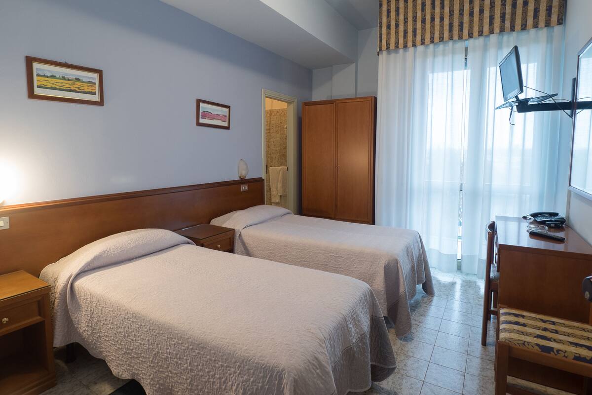 Ein Hotelzimmer mit zwei Betten, einem Holzschrank, einem Schreibtisch mit Stuhl und einem fenster mit Vorhängen.