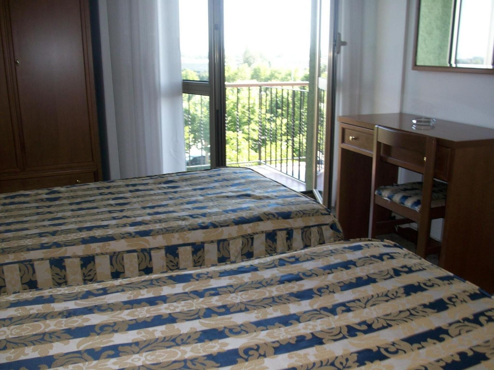 Un dormitorio con cama, un armario de madera, un escritorio pequeño y un balcón con vistas a zonas verdes.