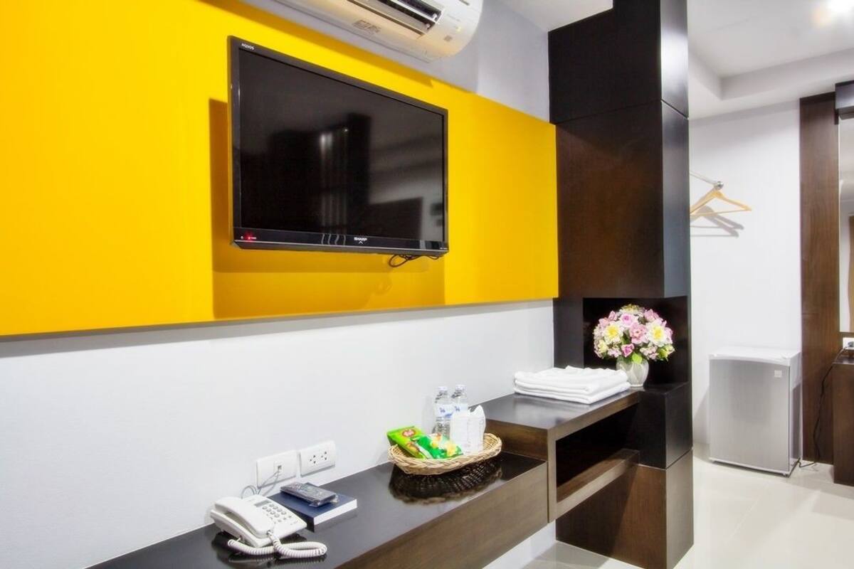 Ein Hotel-Rezeptionsbereich mit einem an einer gelben Wand montierten Flachbildfernseher, einem Empfangstresen mit Telefon, einem Tablett für Toilettenartikel und einer Blumenvase.