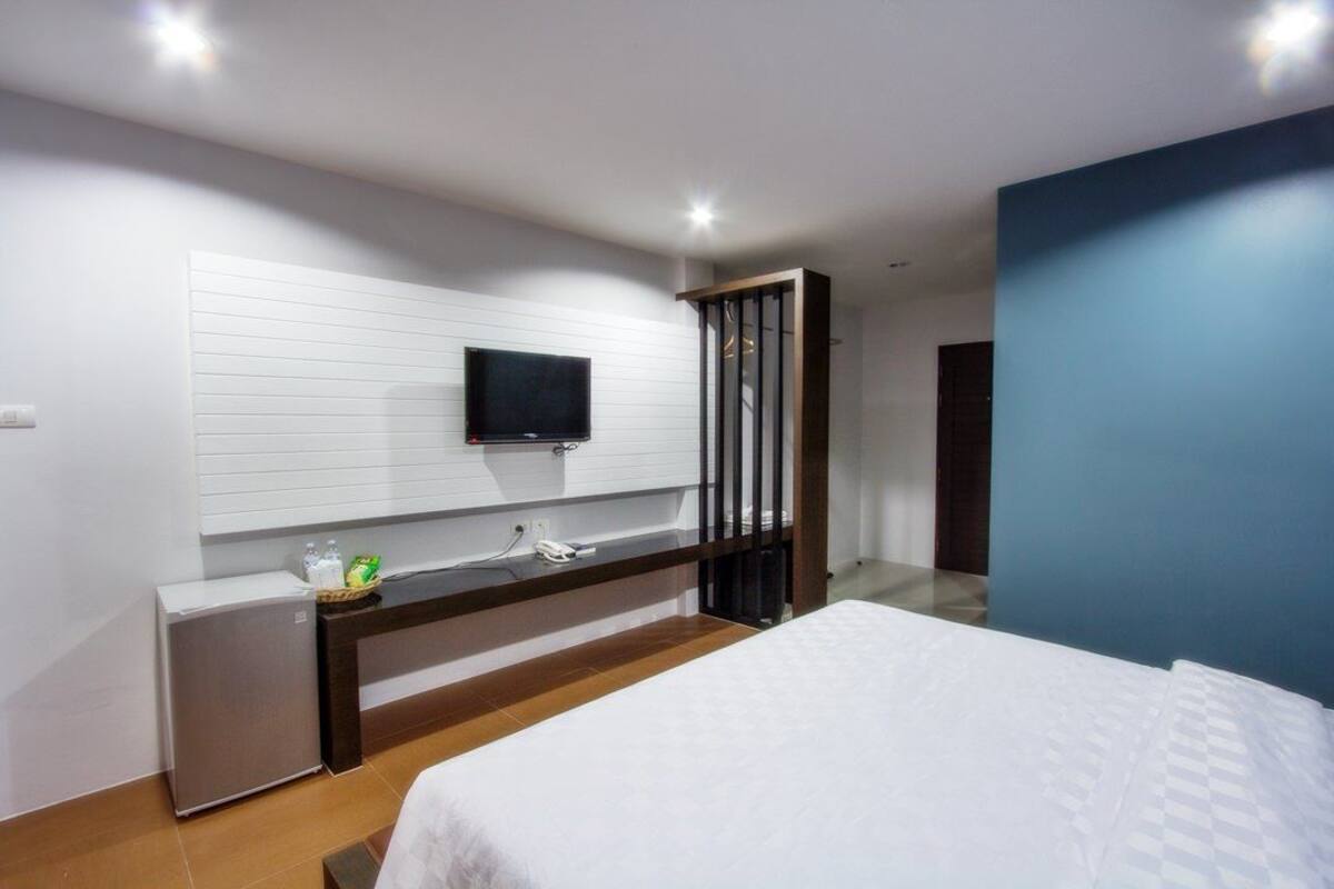 Ein modernes Schlafzimmer mit einem Bett, einem an einer weißen Wand montierten Fernseher, einem kleinen Nachttisch mit Telefon und Flaschen sowie einer blauen Akzentwand.