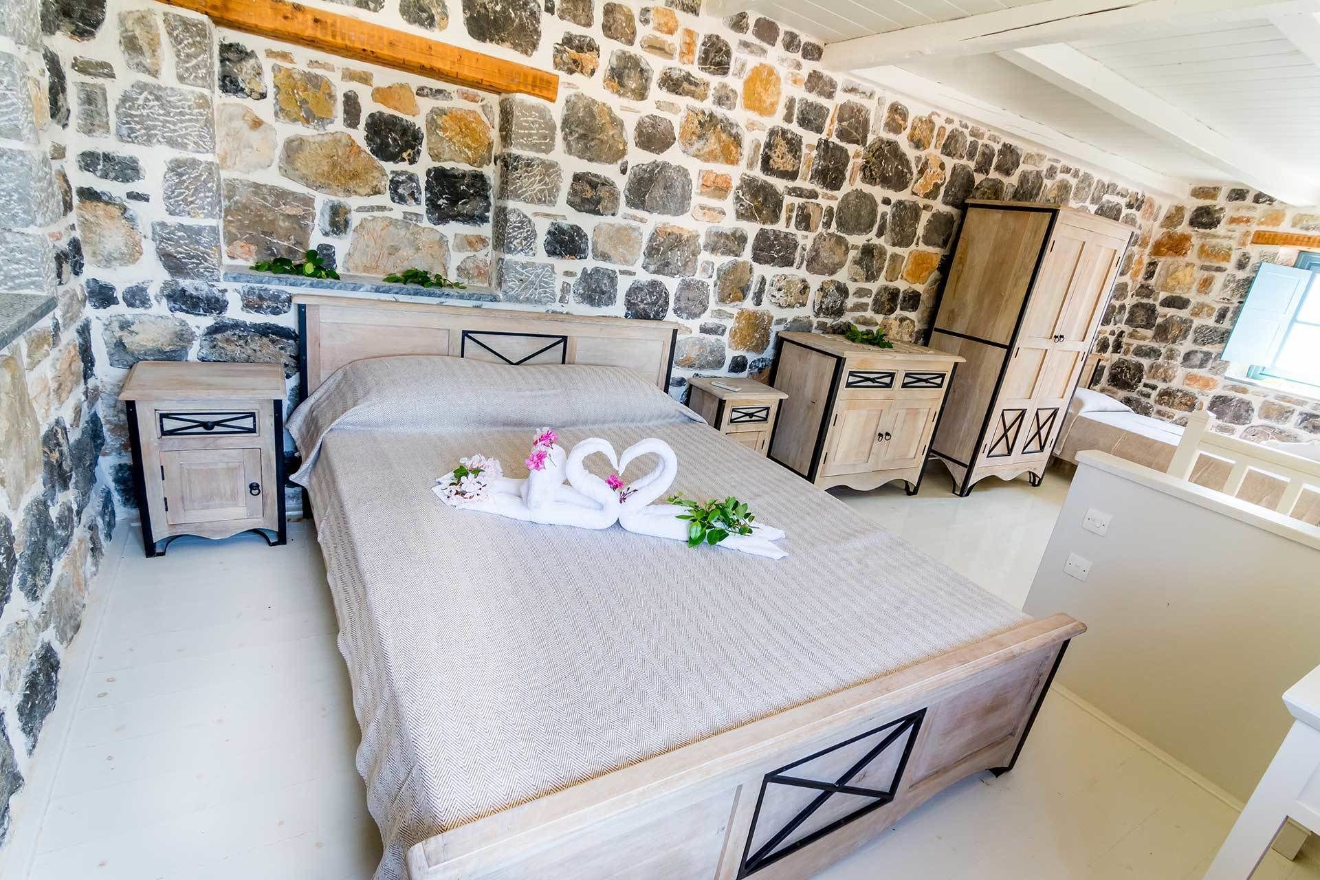 A bedroom with a stone wall, a bed with a heart-shaped towel arrangement, wooden furniture, and a small nightstand.