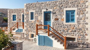 A stone building with blue shutters and wooden railings, featuring a staircase and a small outdoor seating area.