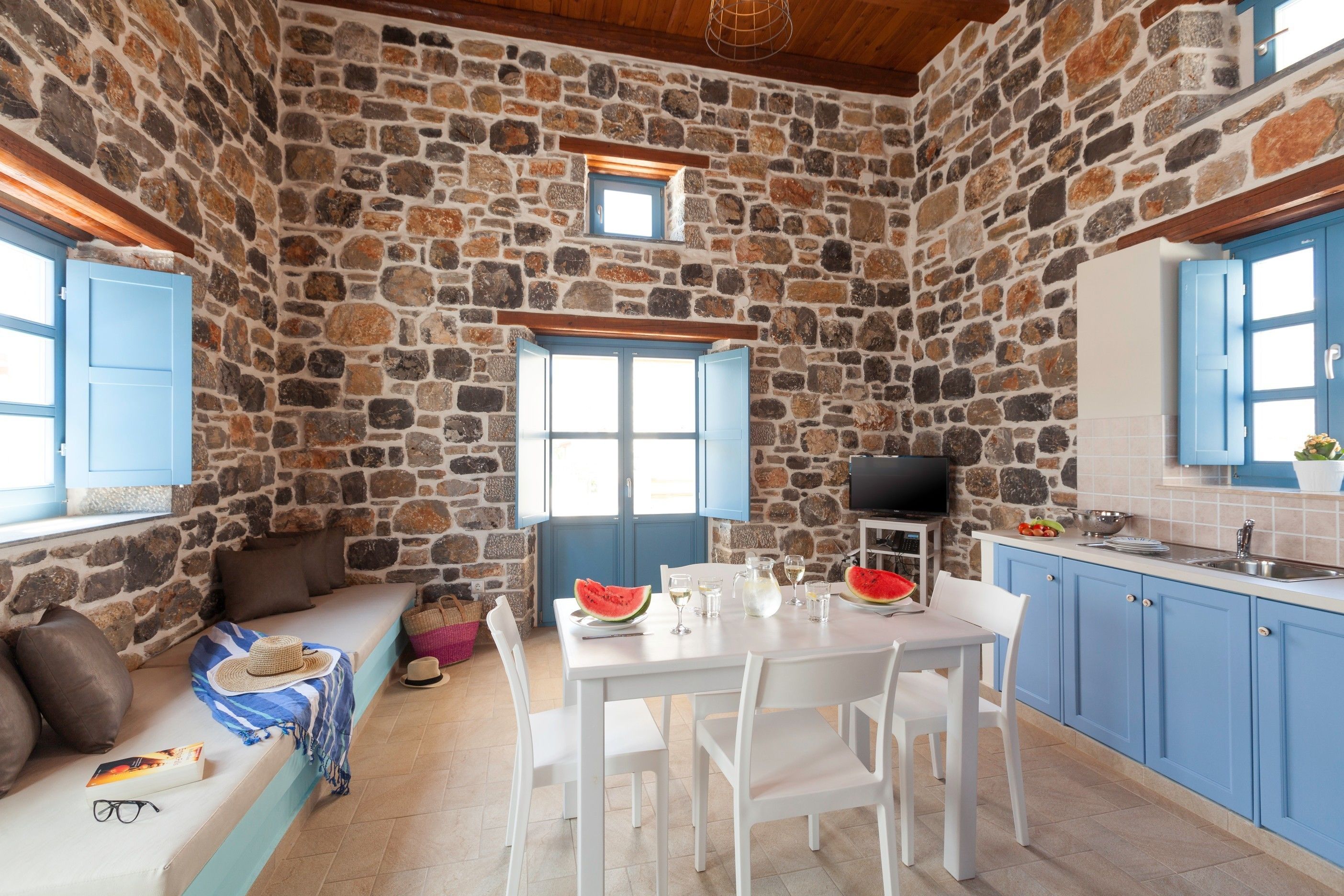 A stone-walled room with a dining table, chairs, a small television, and a window with blue shutters.
