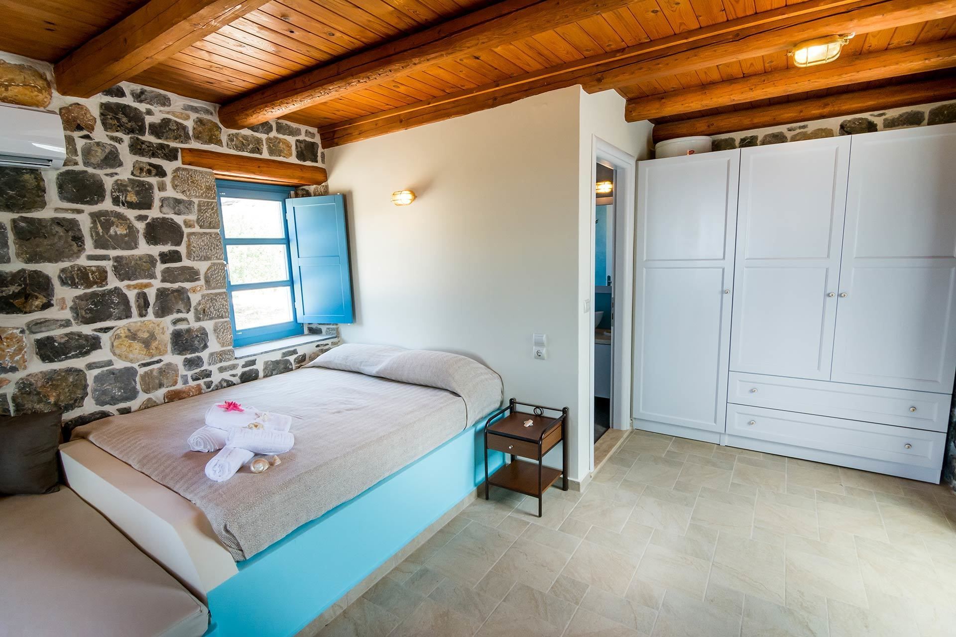 A bedroom with a stone wall, wooden ceiling beams, a bed with a blue headboard, a small bedside table, and a wardrobe.