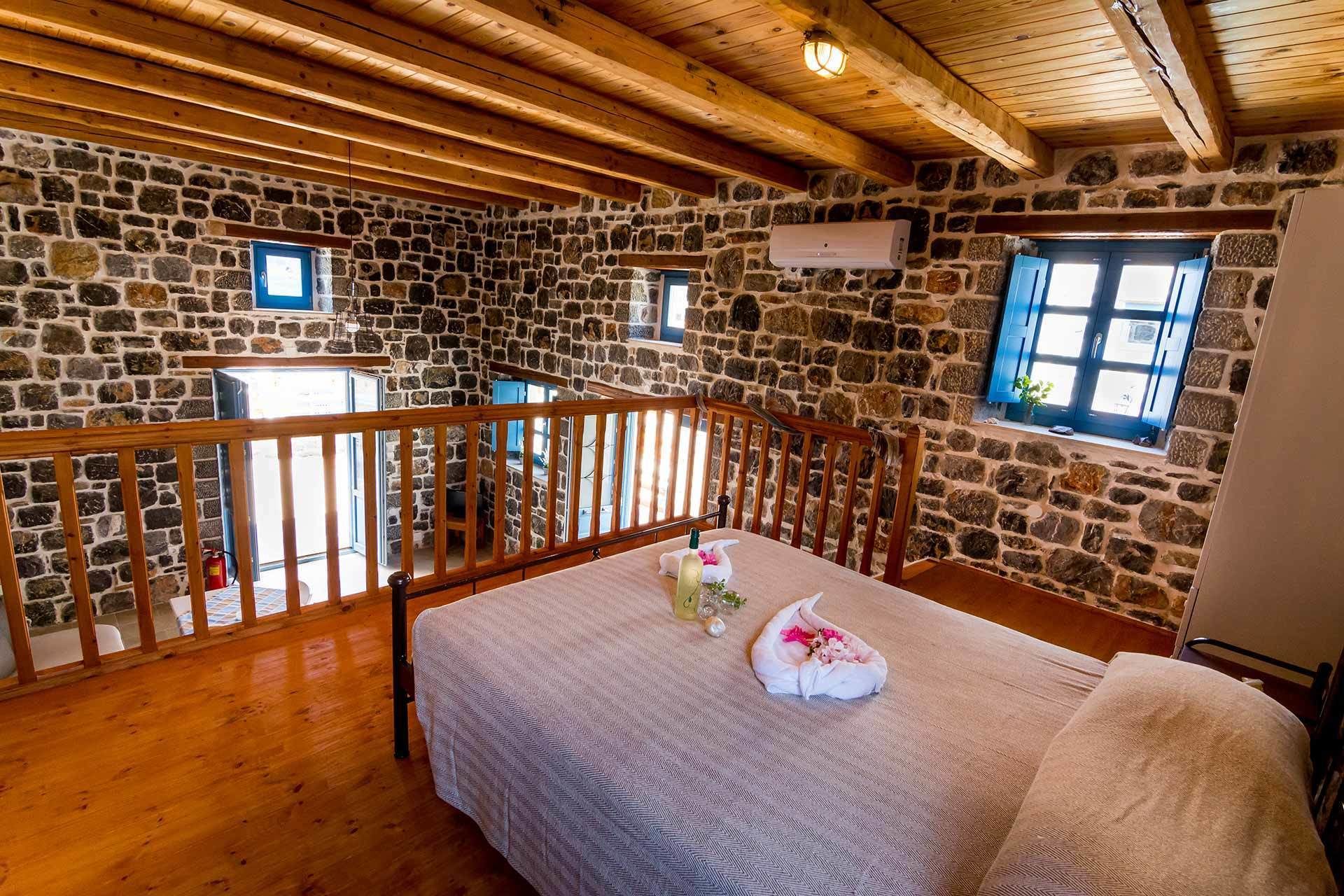 Une pièce aux murs de pierre, avec des poutres apparentes, un balcon, un lit recouvert d’une couverture et une fenêtre munie de volets.