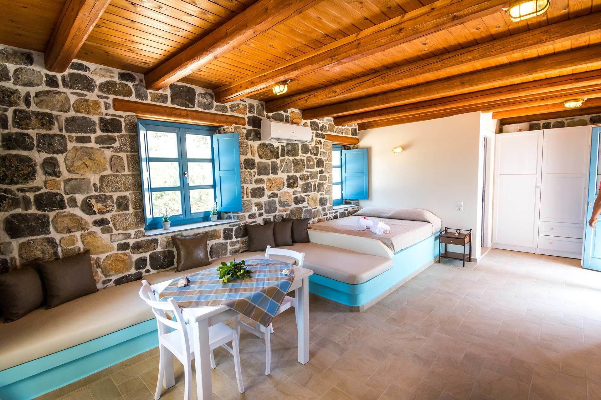 A cozy room with a stone wall, wooden ceiling, a bed with blue bedding, a small table with chairs, and a bench.