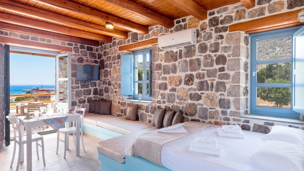 A room with a stone wall, a bed, a table with chairs, and a view of the sea.