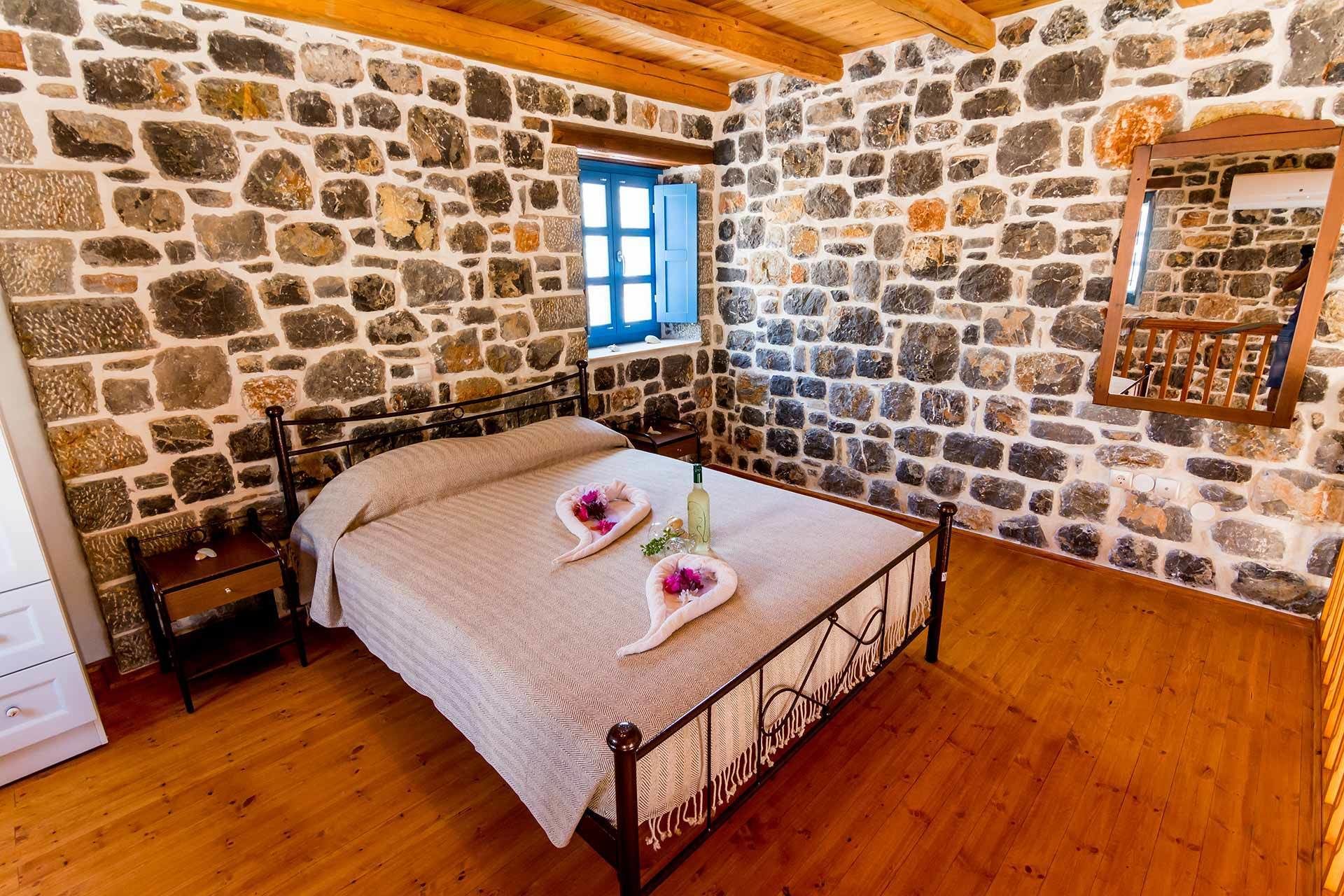A bedroom with a stone wall, a wooden ceiling, a bed with a metal frame, a nightstand, a dresser, and a window with blue shutters.