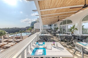 A rooftop terrace with tables and chairs, overlooking a pool and a mountainous landscape.