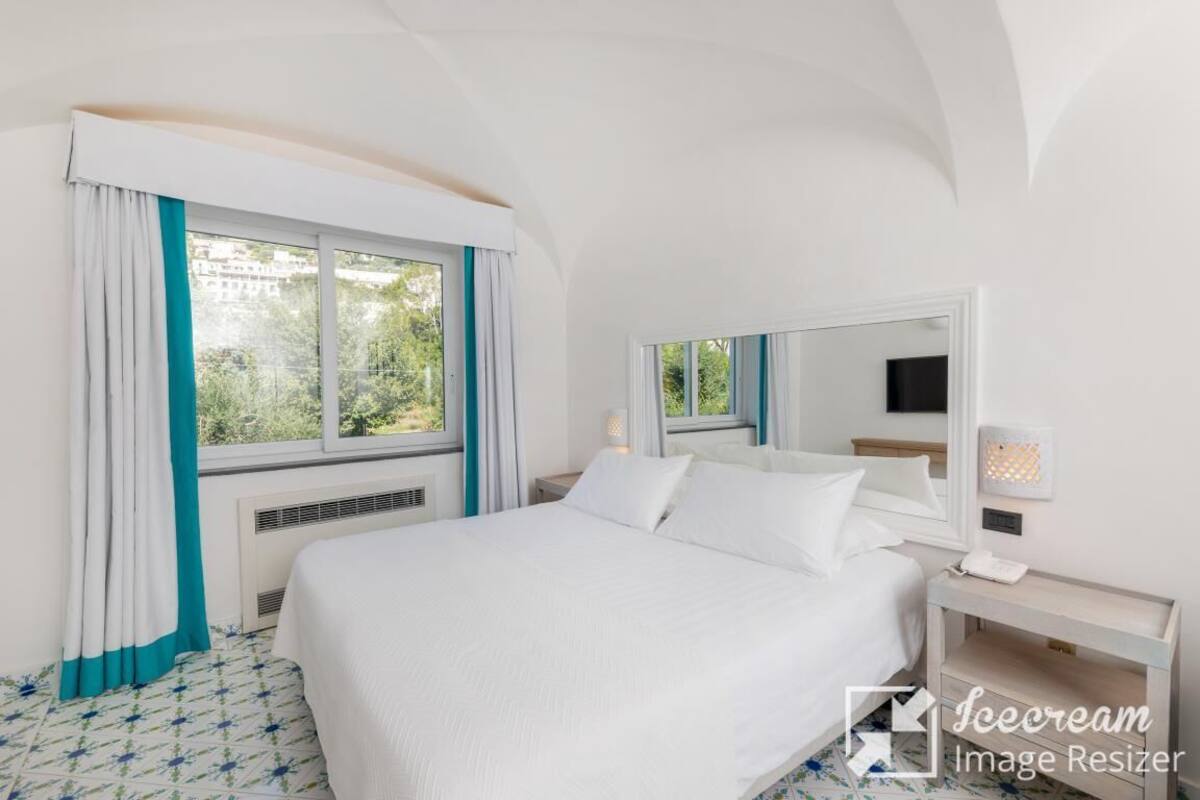 Una habitación de hotel con una cama grande, una mesita de noche con lámpara, una ventana con cortinas y un televisor montado en la pared.