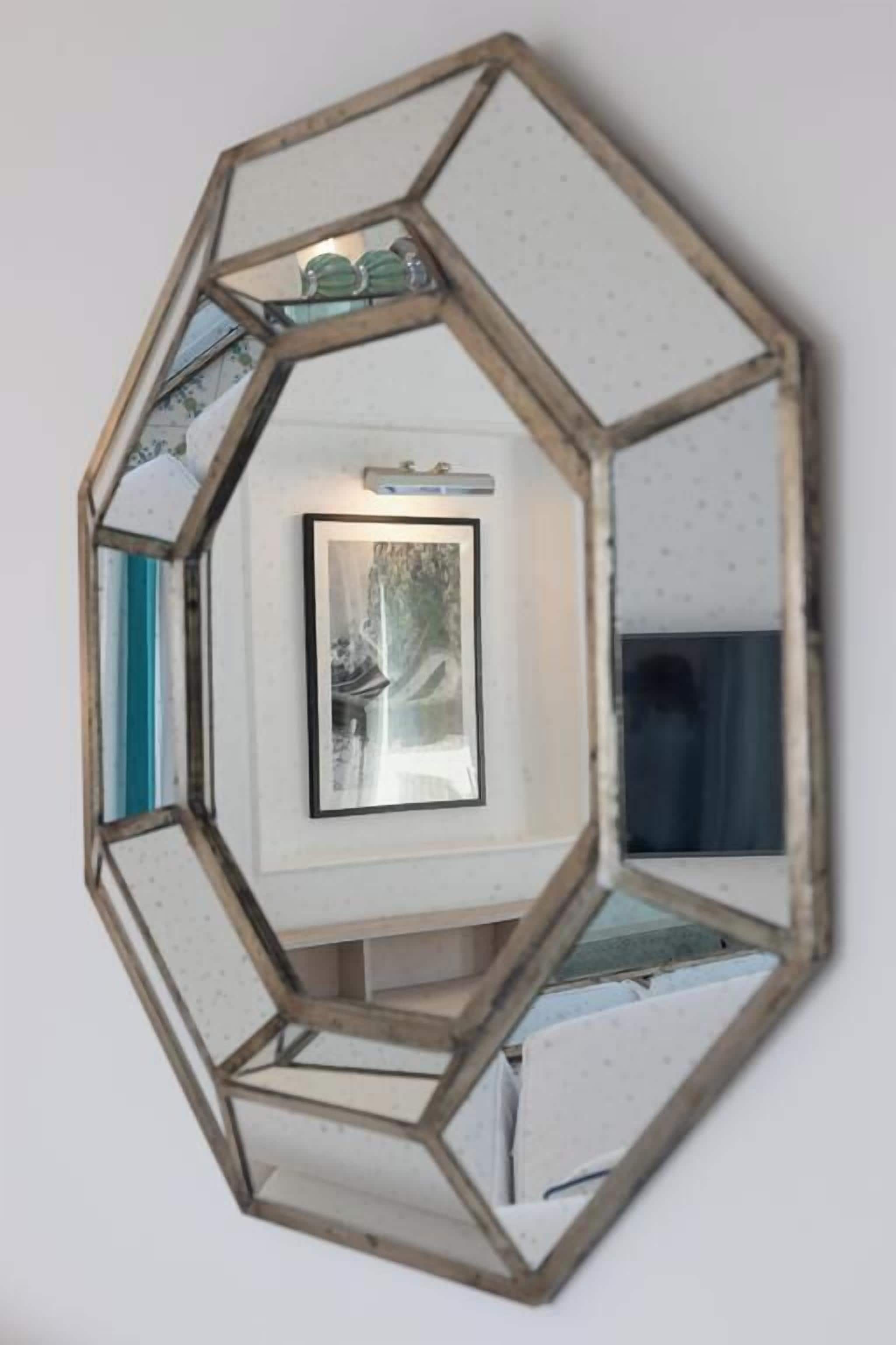 Un miroir hexagonal doté d’un cadre en bois, qui reflète une image encadrée et une étagère ornée d’objets décoratifs.