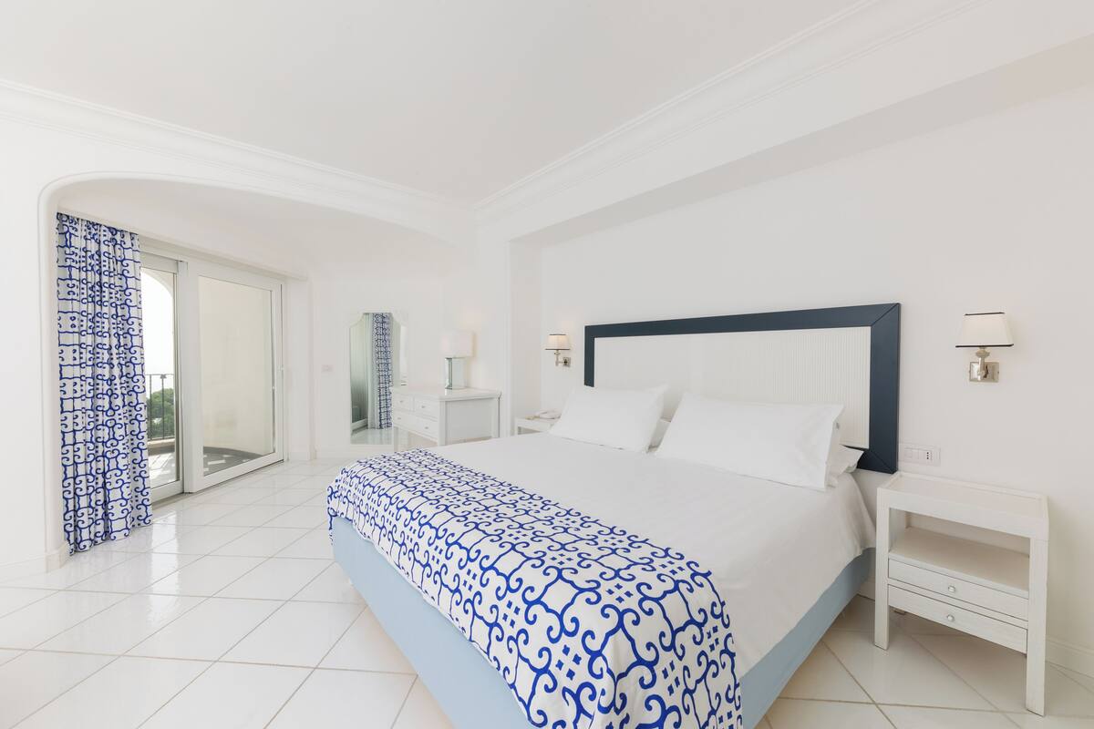 Habitación de hotel con una cama grande, una mesita de noche, una lámpara y vistas al exterior.