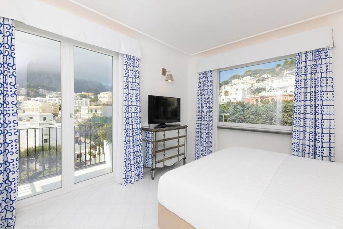 Habitación de hotel con una cama grande, un televisor sobre un soporte y un balcón con cortinas estampadas.